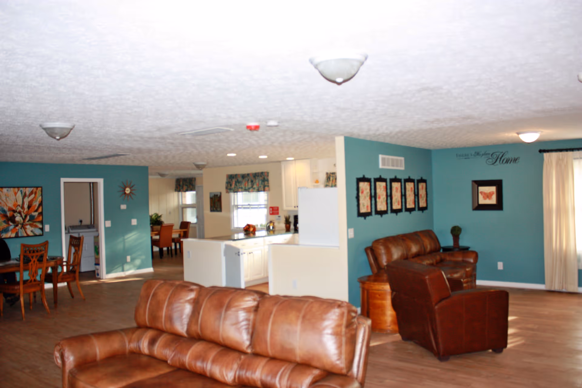 Interior view of a senior living facility common area featuring brown leather sofas and chairs, teal accent walls with framed artwork, a dining area with wooden chairs and table, and a kitchen with white cabinets and floral curtains.
