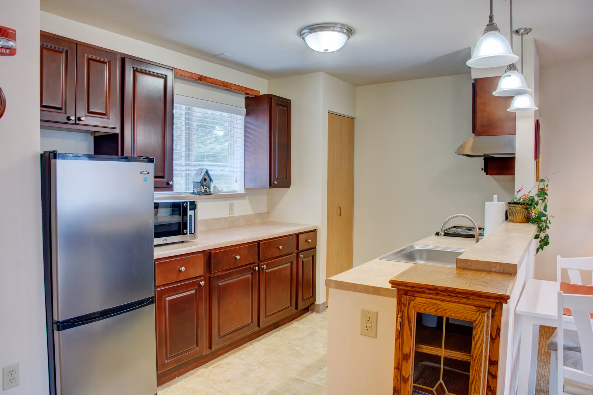 A kitchen with dark wooden cabinets, a stainless steel refrigerator, a microwave on the countertop, a window with blinds, a sink with a faucet on a counter that extends into a breakfast bar with three hanging pendant lights above it, and a small wooden cabinet attached to the side of the counter.