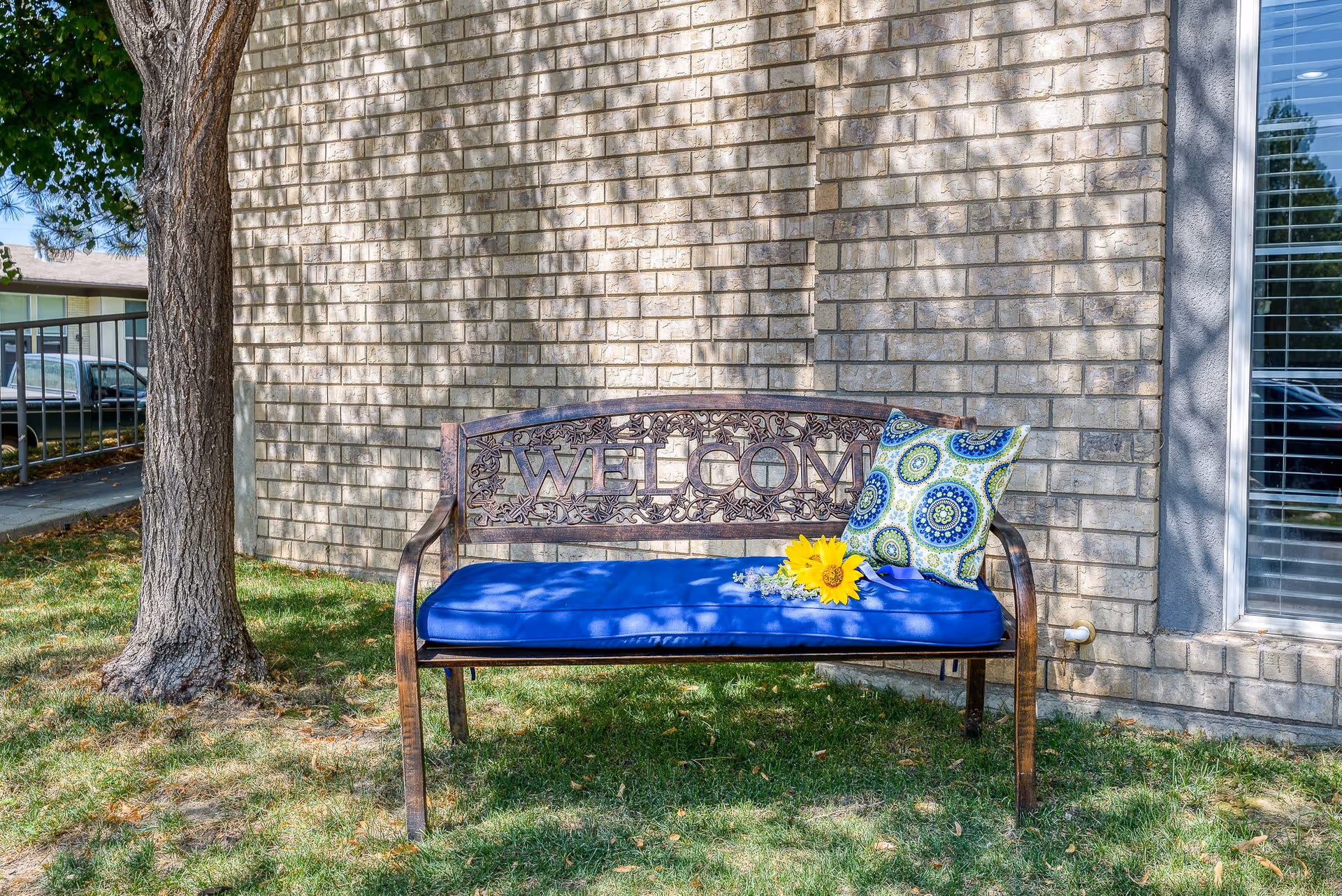 A metal bench with the word 'WELCOME' on the backrest, placed on grass in front of a brick wall. The bench has a blue cushion and a decorative pillow with a blue and green circular pattern. A sunflower and some small flowers are placed on the cushion. A tree trunk is visible to the left, and a window with blinds is on the right side of the image.