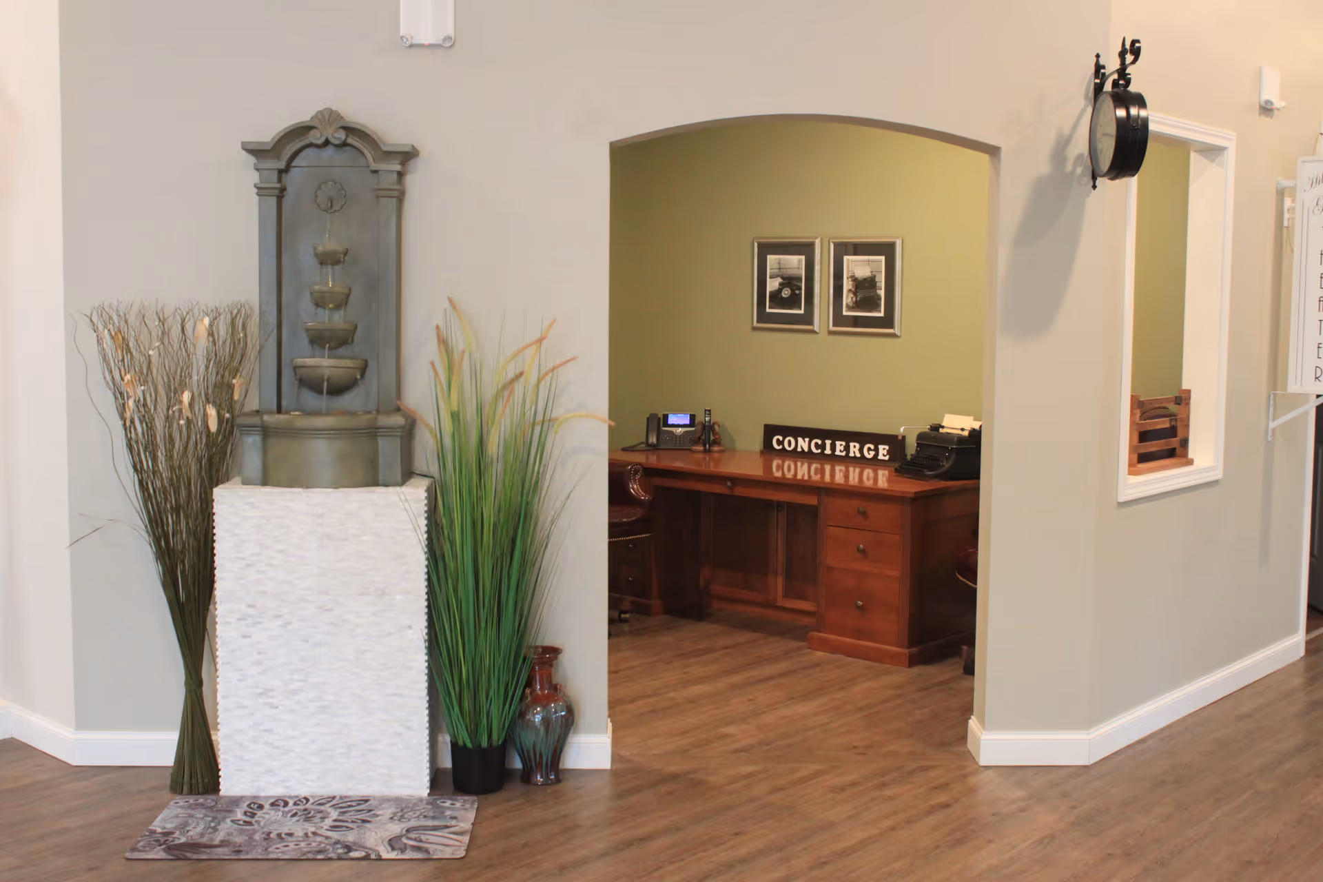 Interior view of a senior living facility showing a small water fountain on a white pedestal with decorative plants on either side. To the right, there is an arched opening leading to a concierge desk with a wooden desk, a sign that reads 'CONCIERGE', two framed pictures on the wall, and office equipment. The floor is wooden, and the walls are painted in neutral tones.