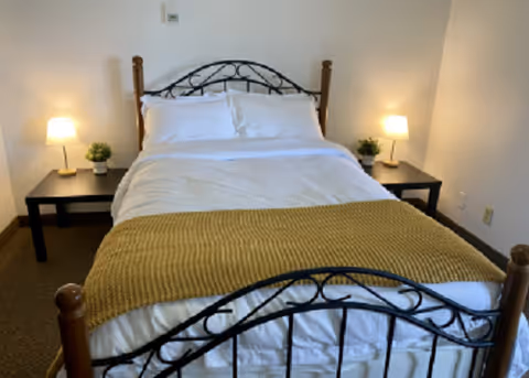 A neatly made bed with white linens and a mustard yellow throw blanket at the foot. The bed has a black metal frame with wooden posts. On each side of the bed, there is a black nightstand with a small lamp and a small green plant. The walls are plain and light-colored.