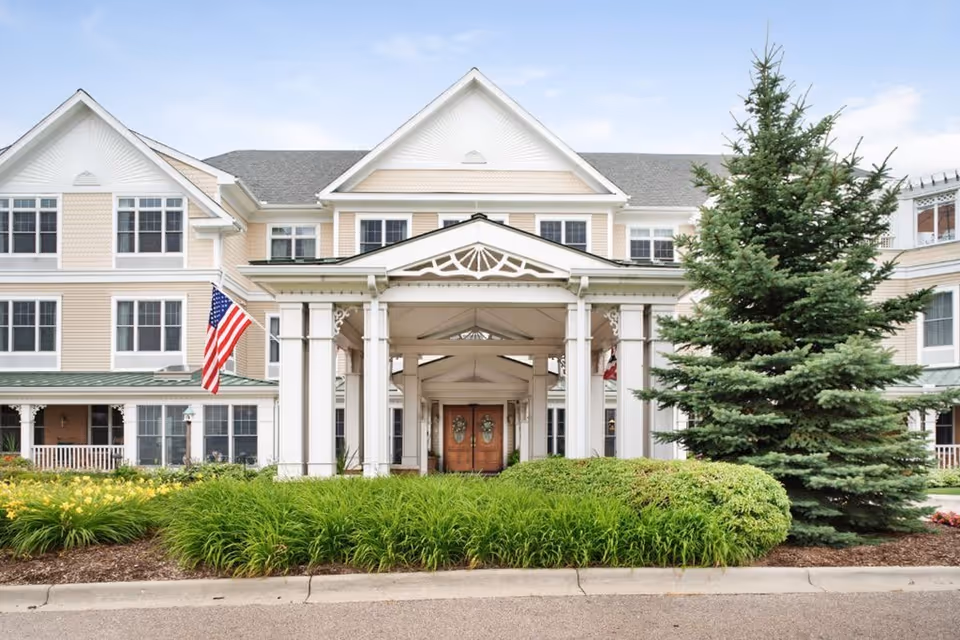 Front exterior view of Sunrise of Shelby Township, a multi-story senior living facility with beige siding, white trim, and a covered entrance supported by white columns. There is an American flag displayed near the entrance and well-maintained landscaping with green bushes and a large evergreen tree.