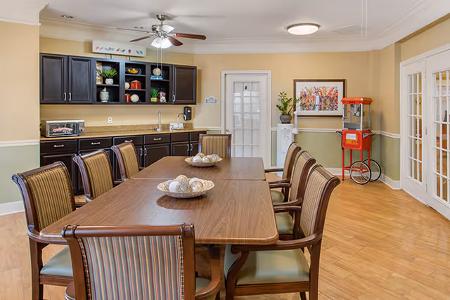 A dining room with a long wooden table surrounded by eight striped cushioned chairs. The room has light green and beige walls, a ceiling fan with lights, and a kitchenette area with dark cabinets, a microwave, and a sink. There is a red popcorn machine in the corner, a water cooler, and a colorful framed artwork on the wall.