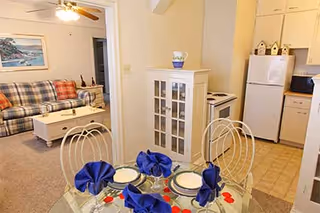 Dining area with a glass-top table set for four in the foreground, a living room with a plaid sofa visible through a doorway and a small kitchenette to the right.
