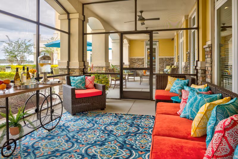 A bright and colorful outdoor patio area at Sonata Windermere featuring a red cushioned wicker sofa with multiple patterned pillows, a matching armchair, and a blue patterned rug. There is a wooden cart with bottles, glasses, and a water dispenser on the left side. The patio is covered with arches and overlooks a garden with trees and umbrellas.