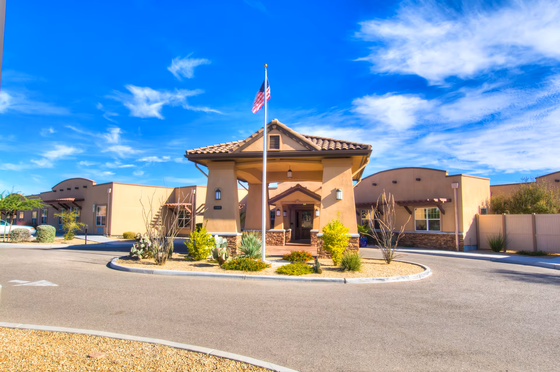 Front exterior view of Canyon Valley Memory Care Residence building with a circular driveway, landscaped plants, and an American flag on a flagpole under a bright blue sky with scattered clouds.