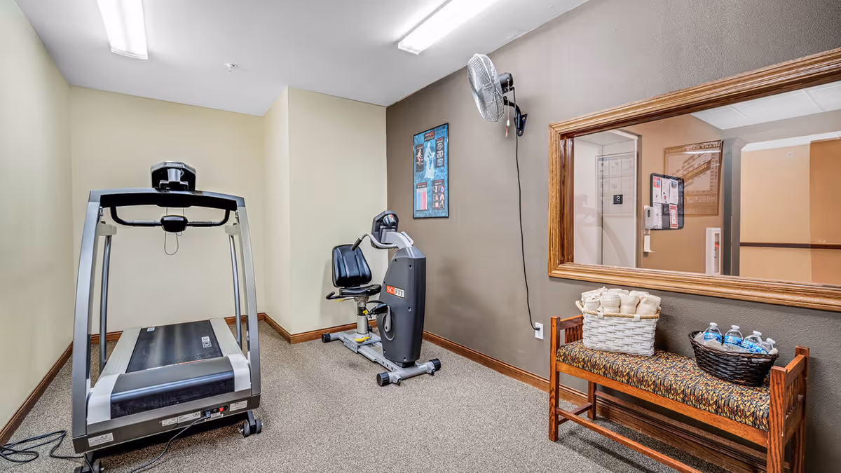 Small indoor exercise room with a treadmill, recumbent bike, bench holding baskets of towels and bottled water, and a window into a hallway.