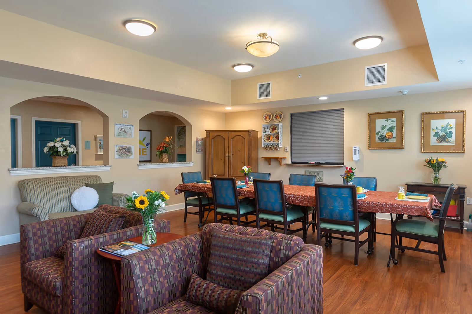 A cozy common area with a long dining table covered with a red patterned tablecloth and surrounded by blue cushioned chairs. In the foreground, there are two patterned armchairs with a small table holding a vase of sunflowers. The room has wooden flooring, beige walls, and ceiling lights. Decorative plates and framed artwork hang on the walls, and there are flower arrangements placed around the room.