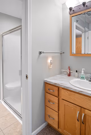 Bathroom with a wooden vanity featuring a marble countertop, a sink, and a mirror cabinet above. There is a towel bar on the wall and a glass-enclosed shower stall with a sliding door. The floor is tiled near the shower and carpeted near the vanity.