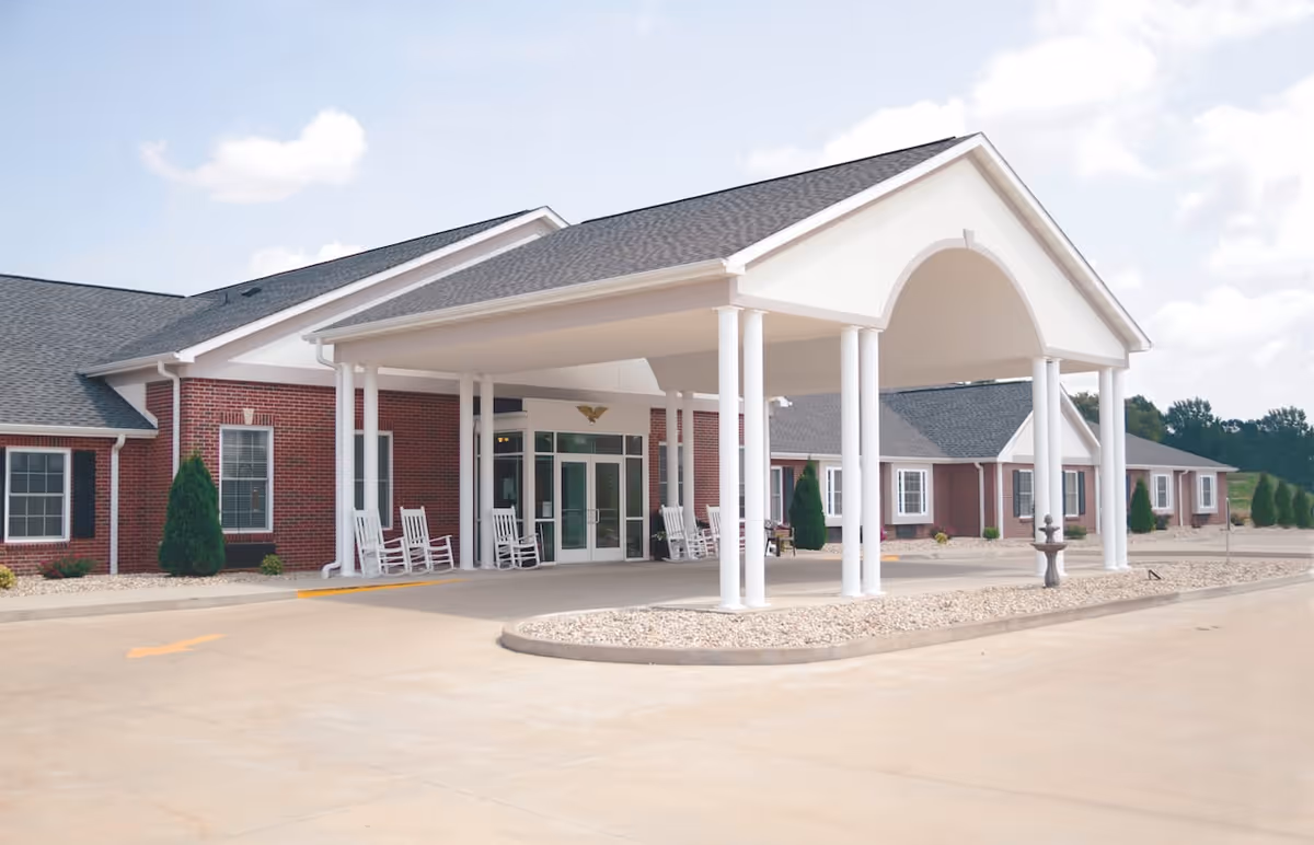 Front exterior view of a single-story brick building with a large covered entrance supported by white columns. Several white rocking chairs are placed under the covered area near the entrance doors. The surrounding area includes a paved driveway and landscaped sections with small bushes and rocks.