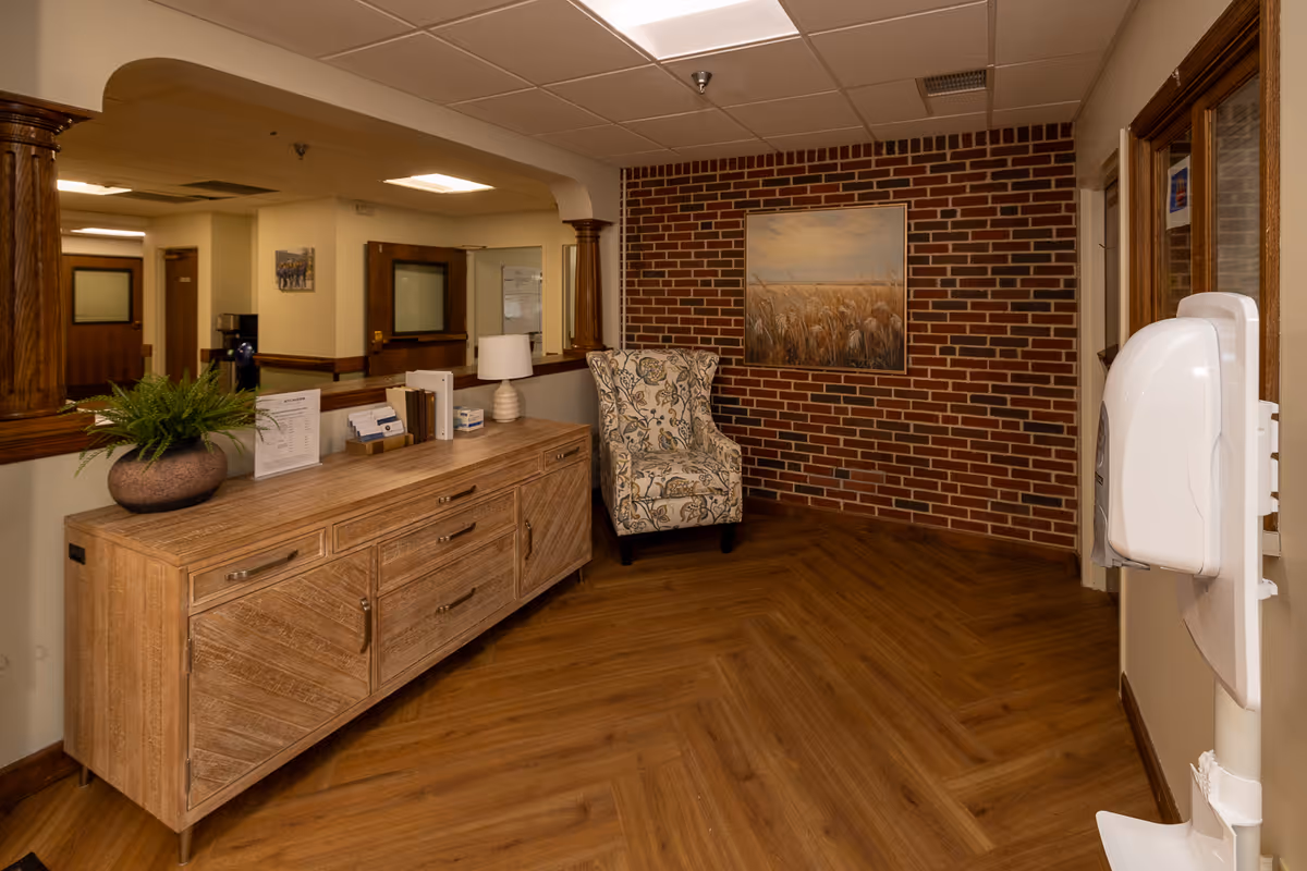 Interior corner of a senior living facility with a wooden sideboard holding a plant, lamp, and informational materials. A patterned armchair is placed against a brick wall with a painting of a wheat field. The floor has a wood-like finish and there is a hand sanitizer dispenser mounted on the wall to the right.