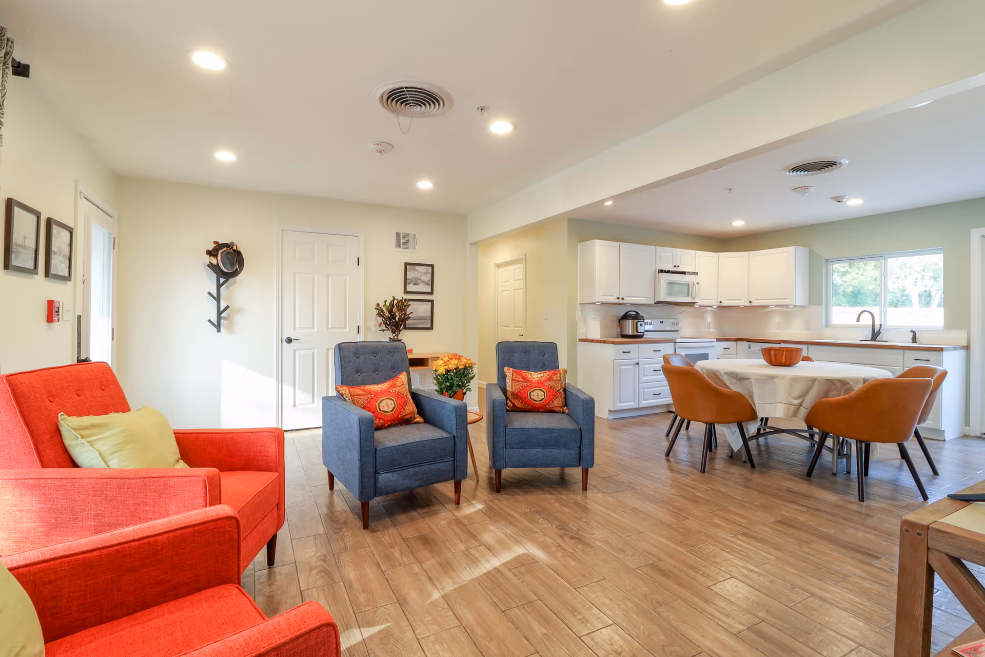 A bright and spacious senior living common area featuring a seating area with two blue armchairs with red patterned pillows and a red armchair with a green pillow. The room has wood flooring and white walls with framed pictures and a coat rack. In the background, there is a kitchen with white cabinets, a microwave, a stove, and a round dining table with four brown chairs. Large windows allow natural light to fill the space.