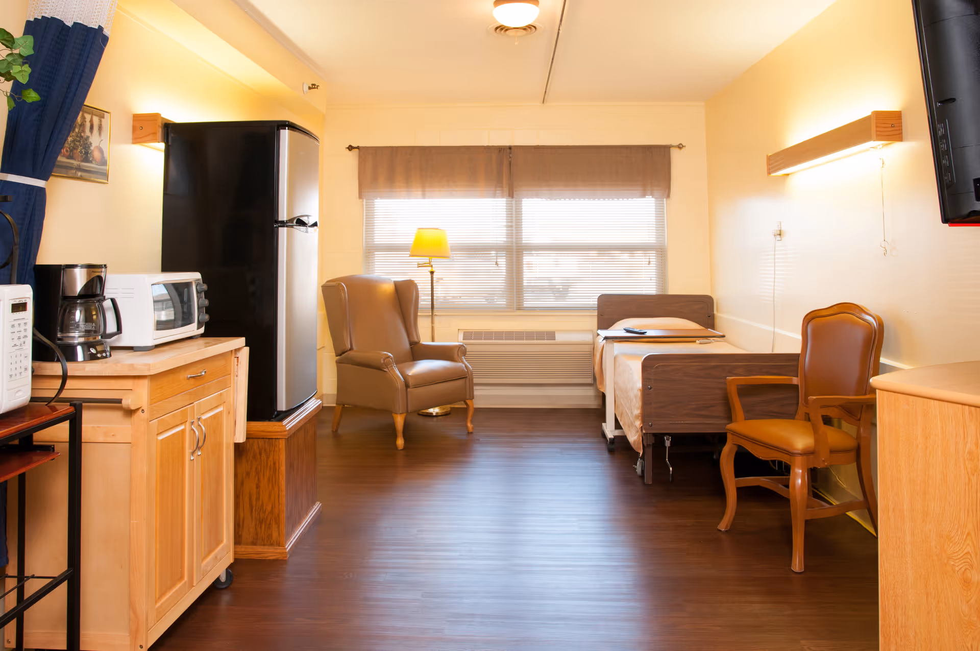 Small senior living studio room with a bed, armchairs, kitchenette with a refrigerator and microwave, and a window.