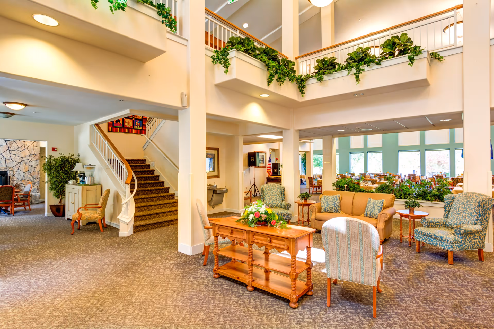 Bright and spacious senior living facility common area with comfortable seating including armchairs and a sofa arranged around a wooden coffee table with a floral centerpiece. There is a staircase leading to an upper floor adorned with green plants on the railing. In the background, a dining area with tables and chairs is visible through large windows letting in natural light.