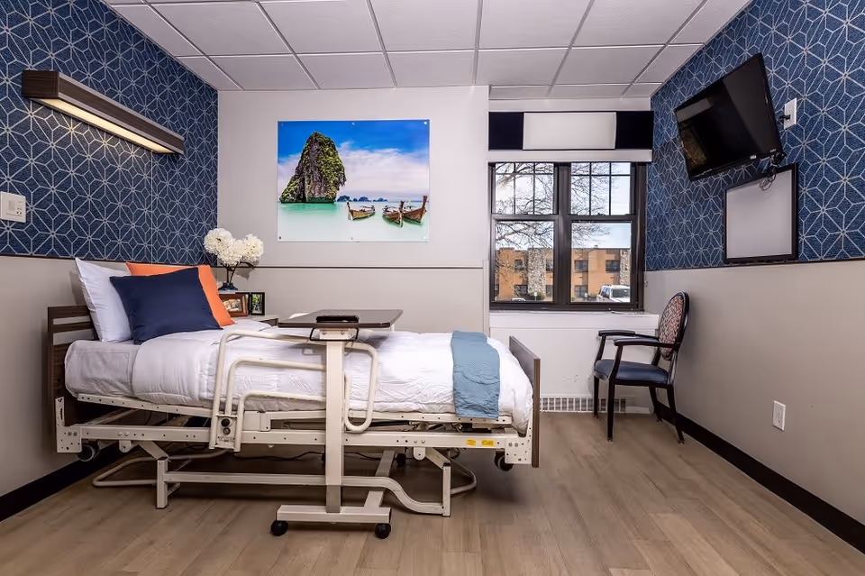 A neatly arranged nursing facility room with a hospital bed made with white linens, blue and orange pillows, and a blue throw blanket. There is a small overbed table, a chair near the window, a wall-mounted TV, and a large window showing an outside view of a building and trees. The walls have a blue geometric patterned wallpaper and a large photo of a tropical beach scene.