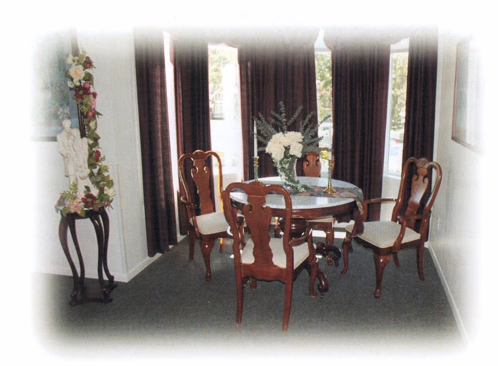 A dining room with a round wooden table surrounded by four wooden chairs with cushioned seats. The table is decorated with a floral centerpiece and two candlesticks. Dark curtains cover the windows in the background, and a small decorative stand with flowers and a statue is positioned to the left.
