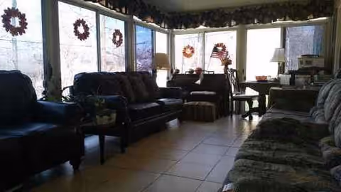 Sunlit communal living room with multiple sofas and chairs facing large windows decorated with wreaths.