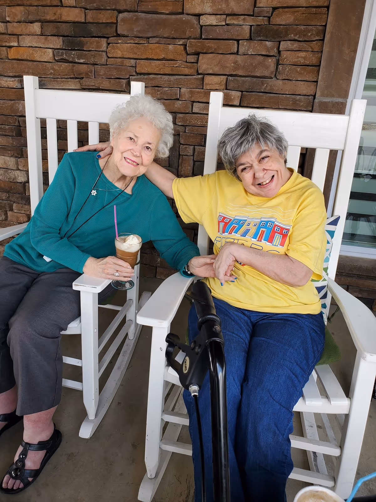 Two elderly women sitting on white rocking chairs on a porch with a stone wall background. One woman in a teal sweater holds a glass with a drink topped with whipped cream and a pink straw, smiling and leaning towards the other woman in a yellow t-shirt and blue pants, who is also smiling and holding hands with her. A black walker is visible in front of the woman in the yellow t-shirt.