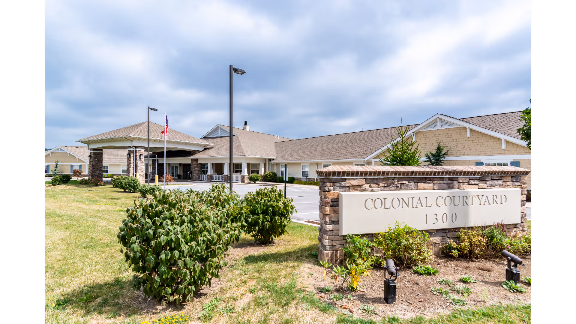 Exterior view of Colonial Courtyard at Clearfield, showing a single-story building with a covered entrance, stone pillars, and an American flag. In the foreground, there is a stone sign with the facility name and address, surrounded by landscaping and bushes under a partly cloudy sky.
