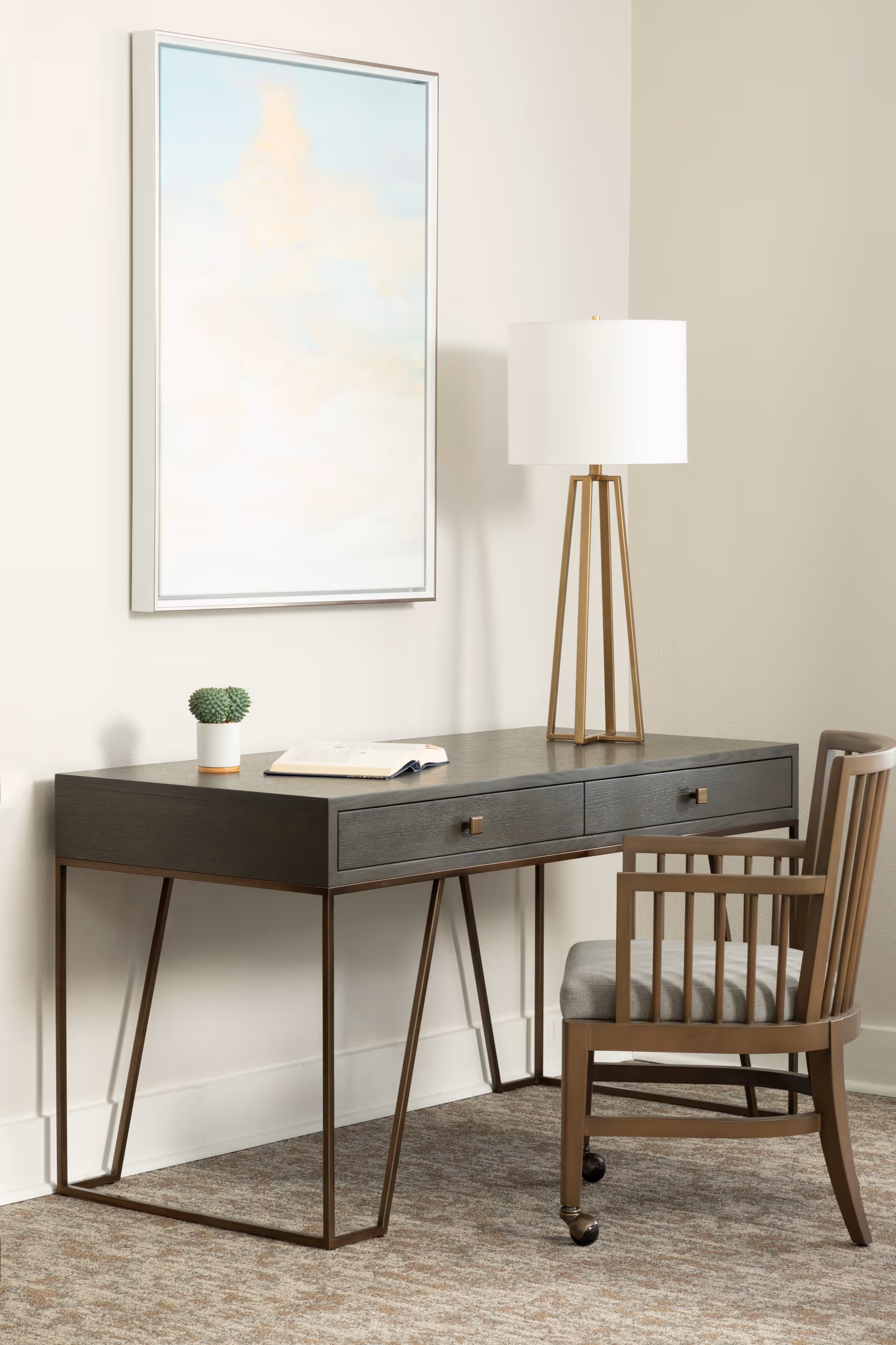 A modern workspace featuring a dark wood desk with two drawers, a small potted cactus, an open book, and a tall lamp with a white shade. A wooden chair with a cushioned seat is positioned in front of the desk. A framed abstract painting with soft pastel colors hangs on the wall above the desk.