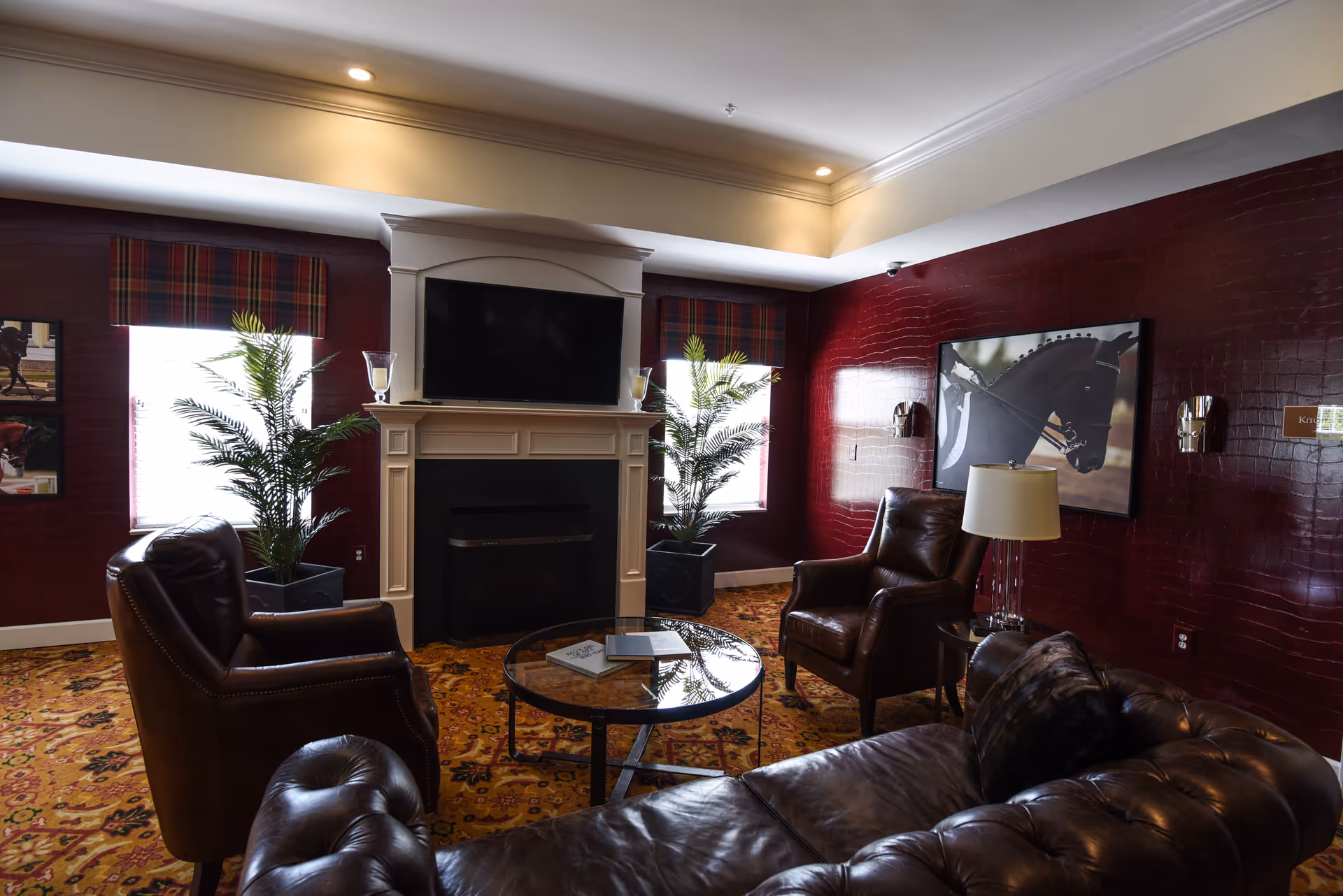 A cozy living room with dark red textured walls, two windows with plaid valances, two potted plants, a white fireplace with a mounted flat-screen TV above it, two brown leather armchairs, a round glass coffee table with books on it, a brown leather sofa, a table lamp, and a framed picture of a horse on the wall.