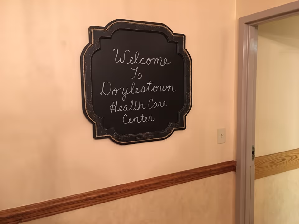 A beige hallway wall with a decorative black chalkboard sign that reads 'Welcome To Doylestown Health Care Center'. The wall has a wooden chair rail and a doorway with a wooden handrail visible inside the adjacent room.