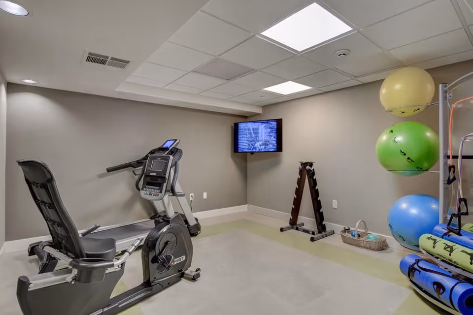 Small fitness room with a recumbent exercise bike, wall-mounted TV, and stacked exercise balls.