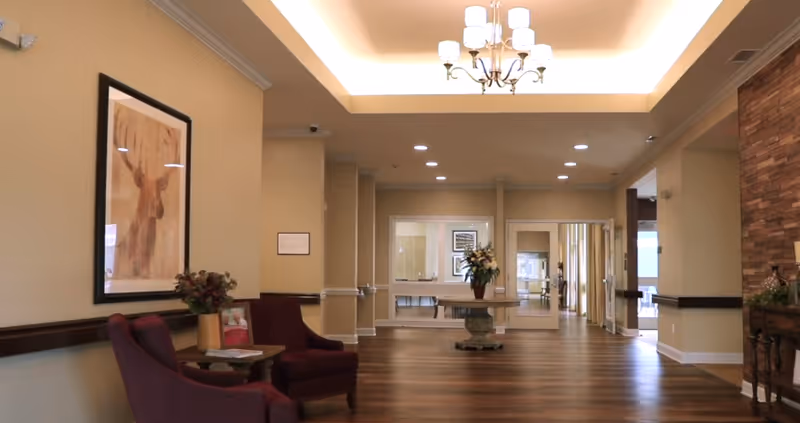 A spacious and well-lit hallway in a senior living facility with wooden flooring, beige walls, and a tray ceiling with a chandelier. There are two purple armchairs and a small table with a flower vase on the left side, a framed picture of a moose on the wall, and a round table with a flower arrangement in the center of the hallway. The hallway leads to other rooms with glass windows and doors.