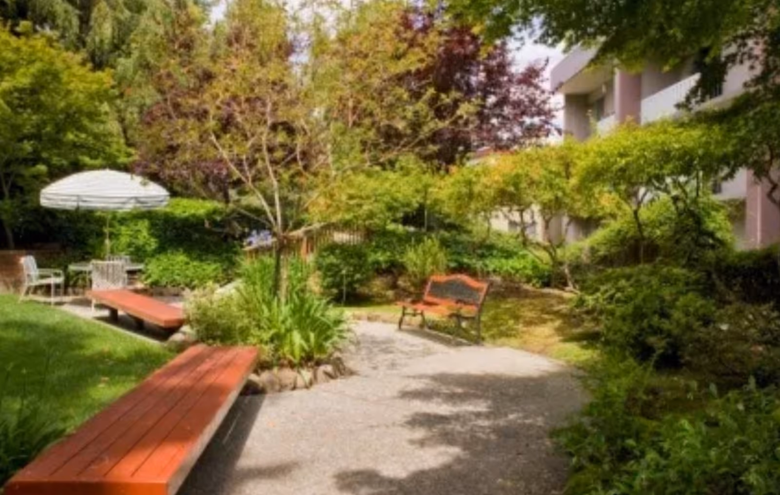 A peaceful outdoor garden area with a paved walking path, red wooden benches, a metal bench, lush green trees and shrubs, and a white patio umbrella with chairs in the background near a building.
