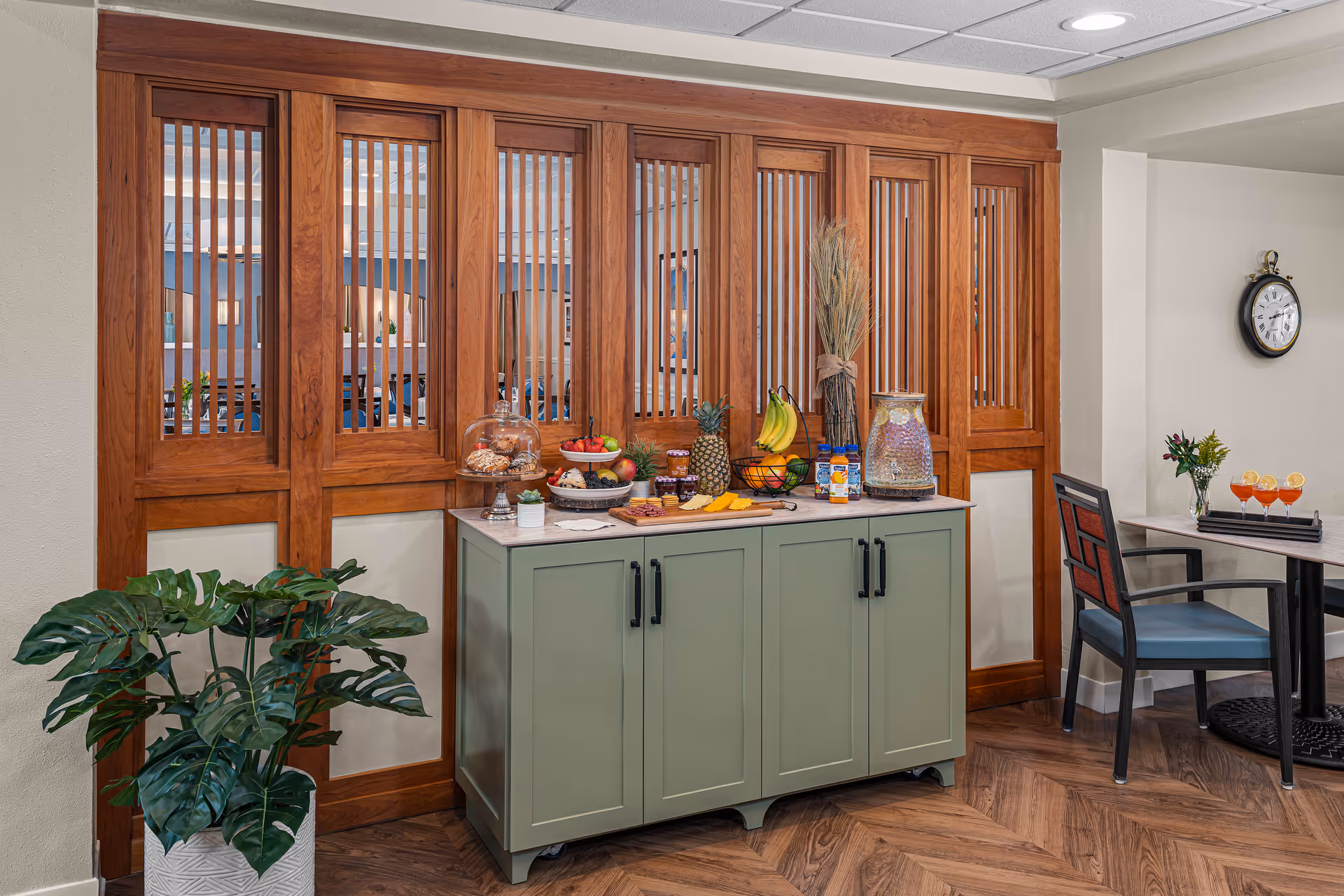 A cozy interior corner featuring a green cabinet topped with a variety of snacks including fruit, cheese, crackers, and jars of preserves. Behind the cabinet is a wooden partition with vertical slats. To the right, there is a small table with a chair, a vase with flowers, and three glasses with orange drinks garnished with lemon slices. A round wall clock is mounted on the wall above the table. A large potted plant is positioned to the left of the cabinet.