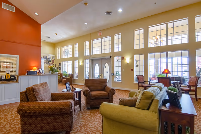 A spacious and well-lit assisted living facility common area with large windows, comfortable armchairs and sofas arranged around small tables, a reception desk with a lamp and decor, and warm yellow and orange walls.
