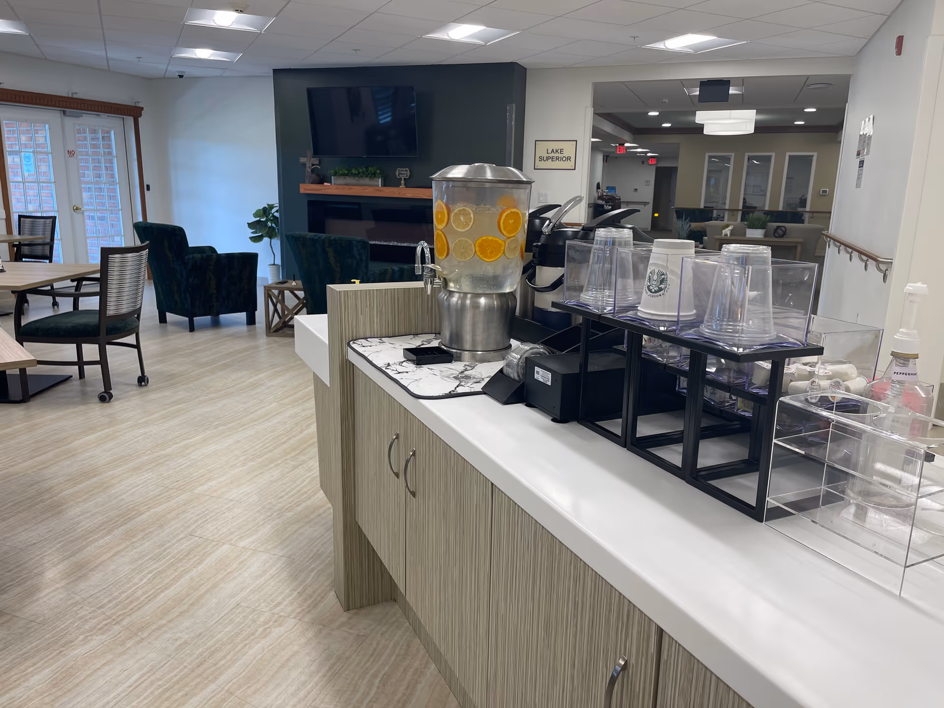 Interior view of a senior living facility common area with a beverage station featuring a dispenser with water and citrus slices, cups, and condiments on a counter. In the background, there are tables and chairs, green upholstered armchairs, a wall-mounted TV above a fireplace, and a sign labeled 'Lake Superior'.