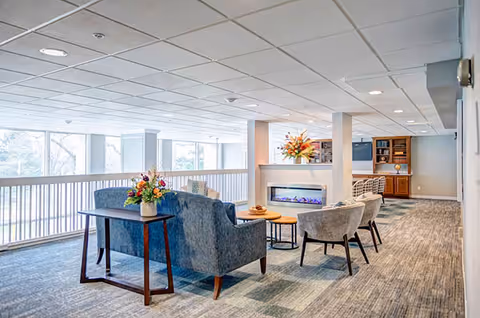 Spacious senior living lounge with upholstered sofas and chairs arranged around a modern electric fireplace and large windows.