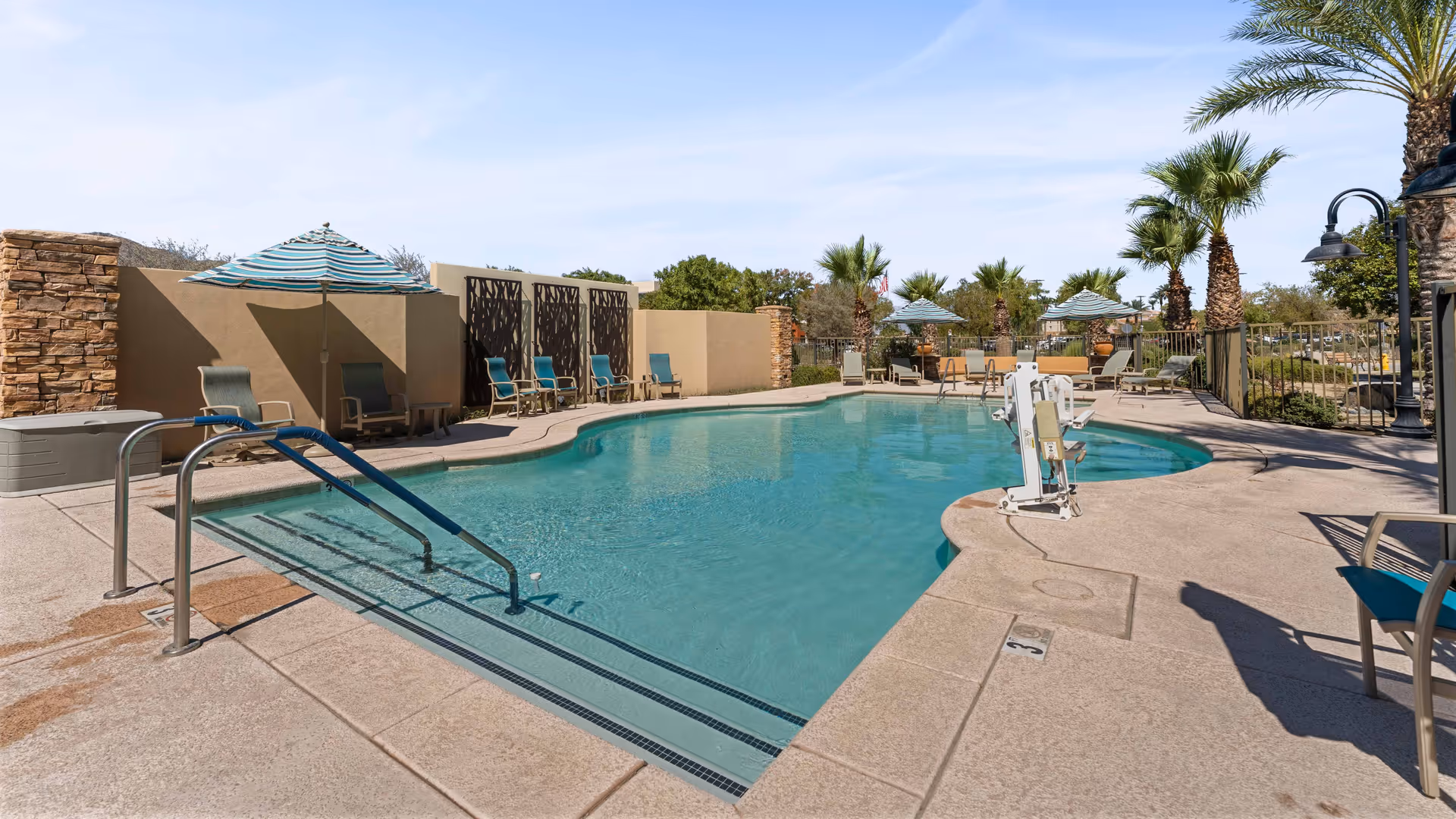 Outdoor swimming pool area with clear blue water, surrounded by lounge chairs and umbrellas. Palm trees and a stone wall are visible in the background under a clear sky.