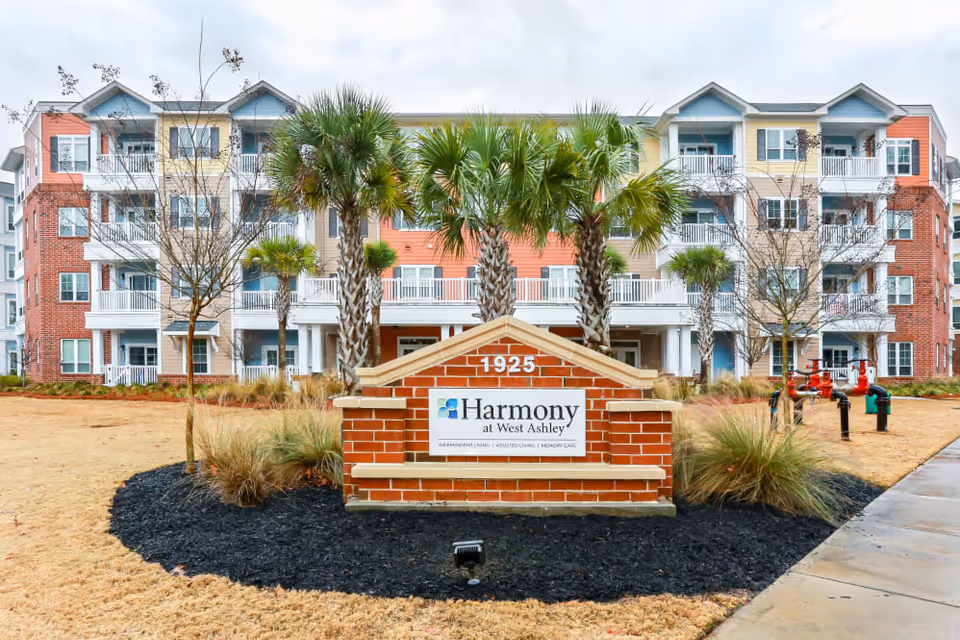Front exterior view of Harmony at West Ashley senior living facility with a brick sign displaying the facility name and address 1925, surrounded by palm trees and landscaping, with a multi-story residential building in the background.