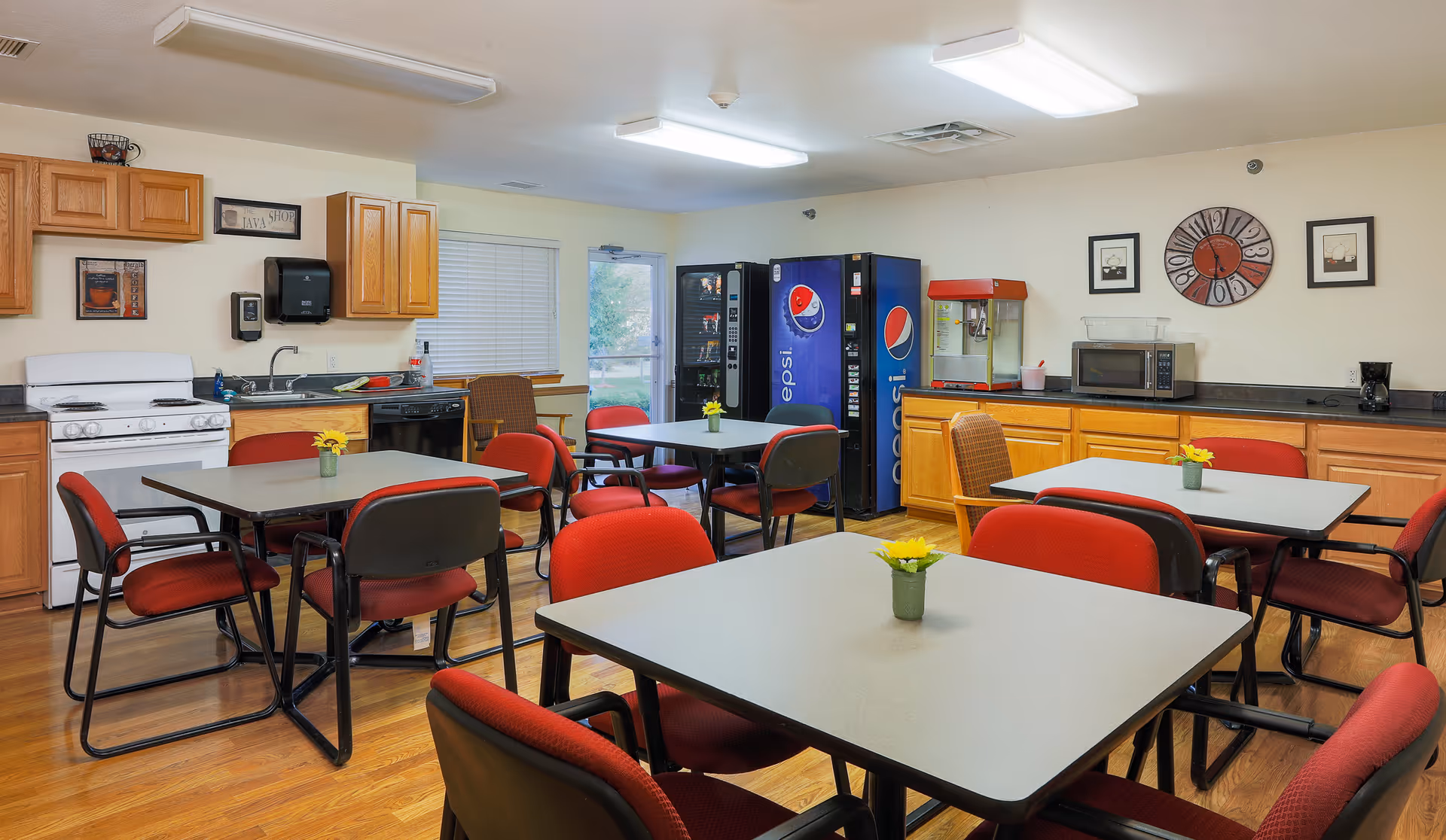 Dining room with multiple tables and red chairs, a kitchenette area, and vending machines.