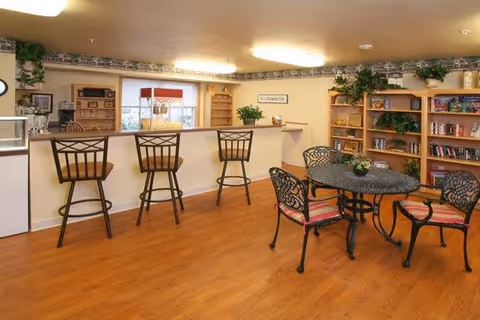 A cozy common area with a wooden floor featuring a round metal table with four chairs that have red and white striped cushions. Behind the table is a counter with three high chairs and a popcorn machine on the counter. The walls have shelves filled with books and decorative plants, and there are additional plants on top of the shelves and counter.