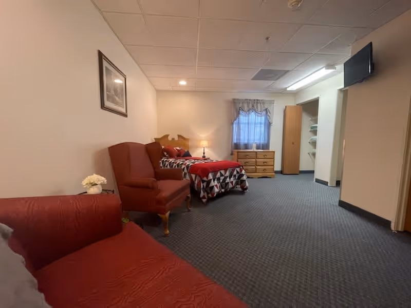 A cozy bedroom in an assisted living facility featuring a single bed with a geometric patterned bedspread and red blanket, a wooden dresser under a window with a curtain, a red upholstered armchair, a red sofa, a small lamp on a nightstand, and a framed picture on the wall.