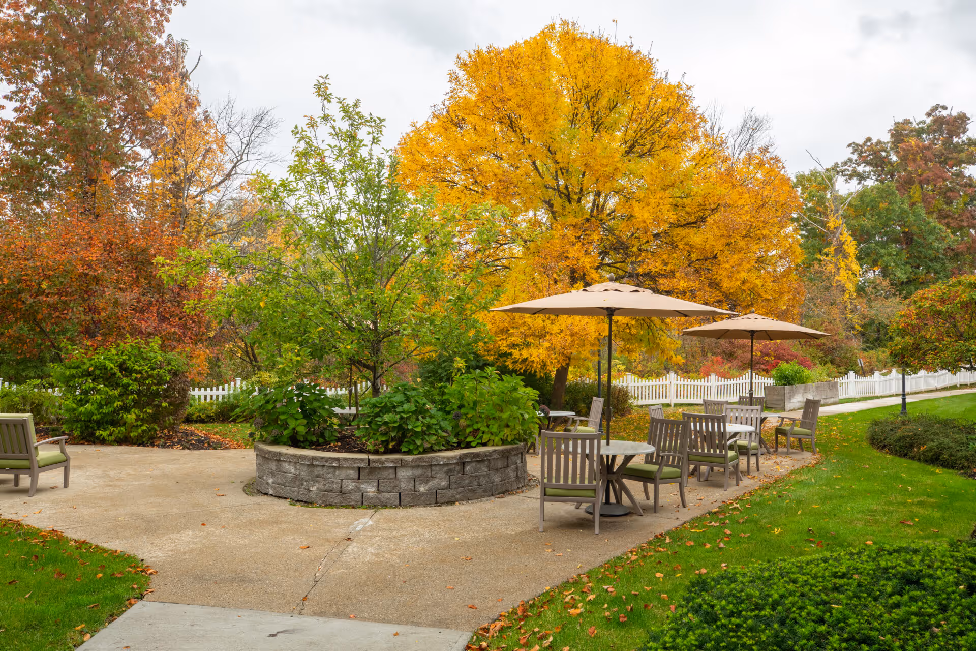 Outdoor patio area with several tables and chairs under beige umbrellas, surrounded by lush greenery and trees with autumn foliage in shades of yellow, orange, and red. A white picket fence runs along the background.