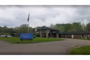 Exterior view of the Embassy of Tunkhannock facility showing a single-story building with a covered entrance, an American flag on a flagpole, a blue sign with the facility name, a parking lot with several cars, and green trees and grass surrounding the area under a cloudy sky.