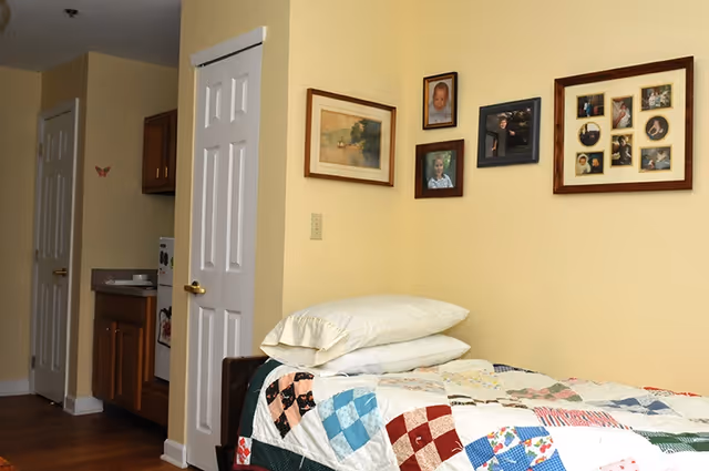 Small bedroom with a single bed covered by a patchwork quilt and stacked pillows, framed photos on a pale yellow wall, and a kitchenette and closet visible in the background.