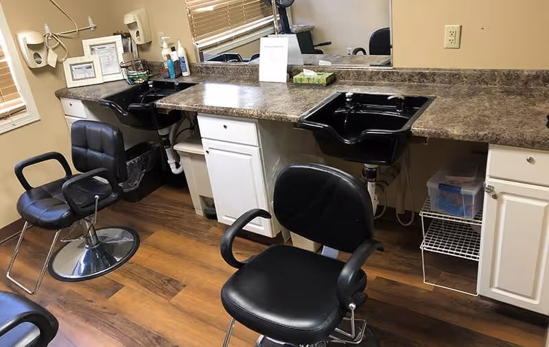 Interior view of a hair salon area with two black salon chairs in front of a countertop with two black sinks. The countertop has various hair care products, a tissue box, and a small sign. There is a large mirror behind the countertop and windows with blinds on the left side. The floor is wooden.
