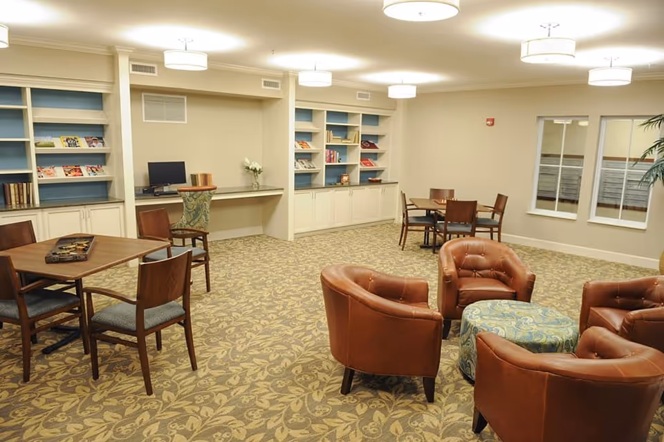 A bright communal lounge with leather armchairs, tables, bookshelves, and a small computer desk.