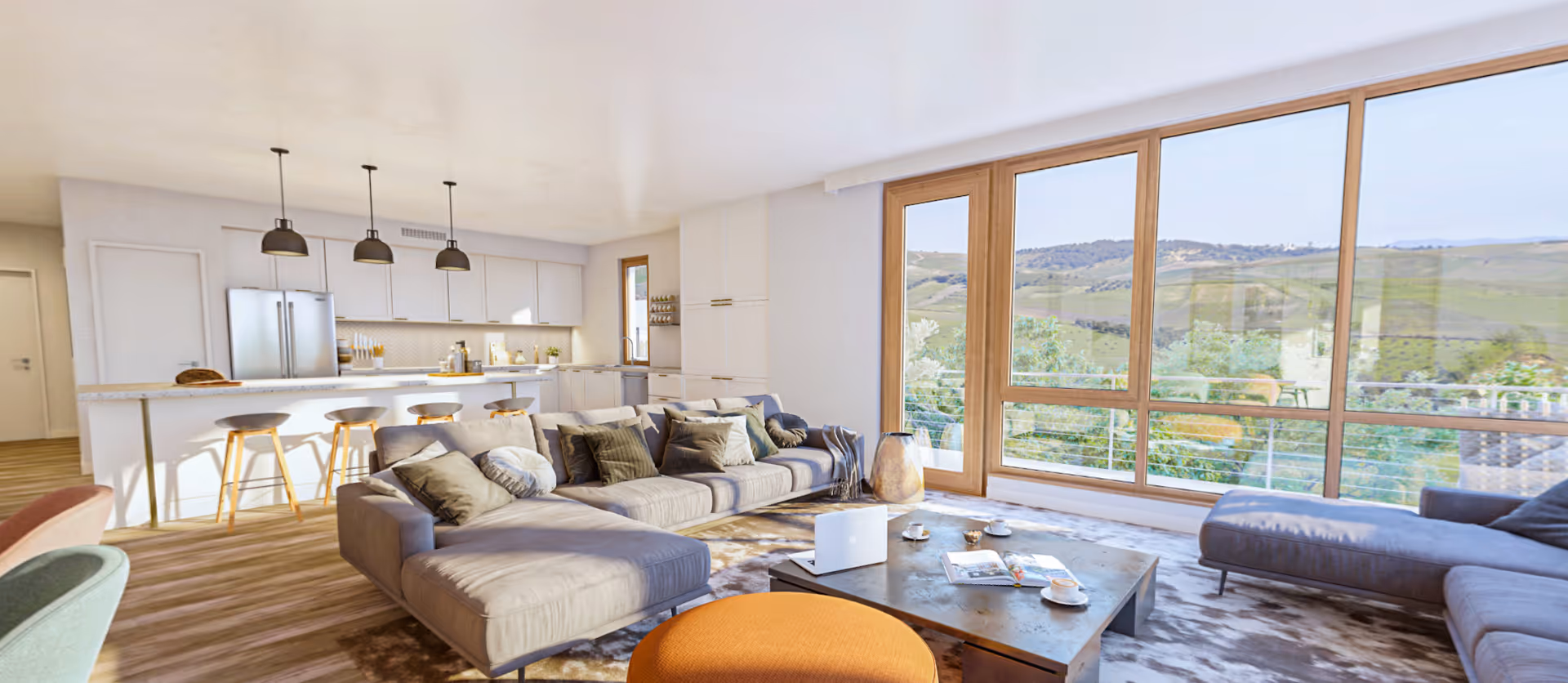 Bright and modern open-concept living room and kitchen area with large windows offering a scenic view of green hills. The living room features a large gray sectional sofa with multiple cushions, a low coffee table with a laptop, cups, and a magazine, and an orange ottoman. The kitchen has white cabinetry, a marble countertop island with four wooden stools, and three pendant lights hanging above.