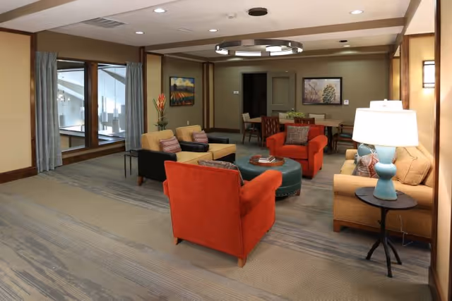 A cozy living room area in a senior living facility with beige walls and carpeted floor. The room features two orange armchairs, two beige armchairs with striped cushions, a beige sofa with patterned pillows, a round green ottoman in the center, a side table with a blue lamp, and framed artwork on the walls. There are large windows with blue curtains on the left side and a dining table with chairs in the background.