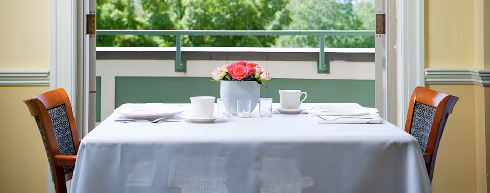 A small dining table set for two with white tablecloth, white cups and saucers, napkins, and a vase of pink and white flowers, positioned near an open door leading to a balcony with green foliage visible outside.