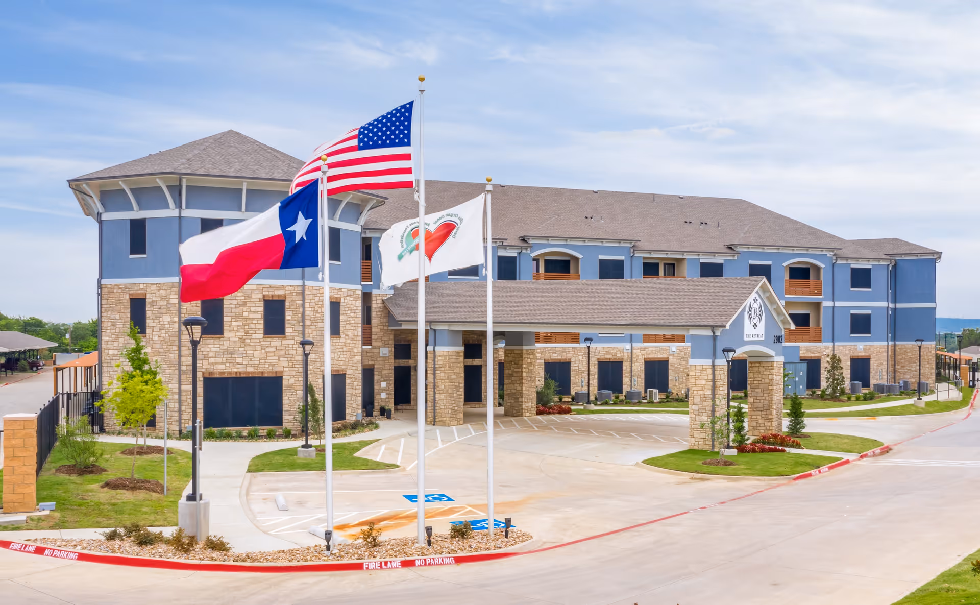 Exterior view of a senior living facility building with a covered entrance and three flagpoles displaying the American flag, Texas state flag, and a facility flag. The building has a stone and blue facade with multiple windows and a well-maintained driveway and landscaping.