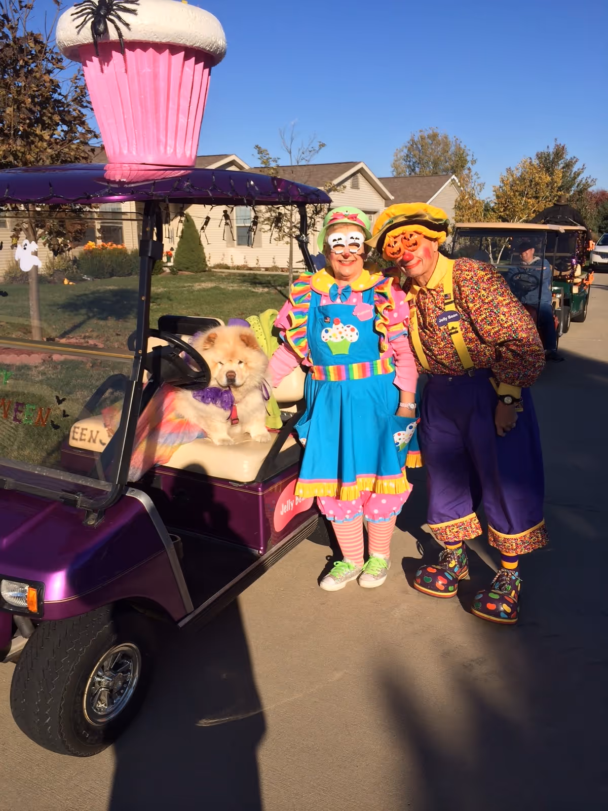 Two people dressed as colorful clowns standing next to a purple golf cart with a large pink cupcake decoration on top. A fluffy dog wearing a purple outfit sits on the golf cart seat. The scene is outdoors on a sunny day with houses and trees in the background.