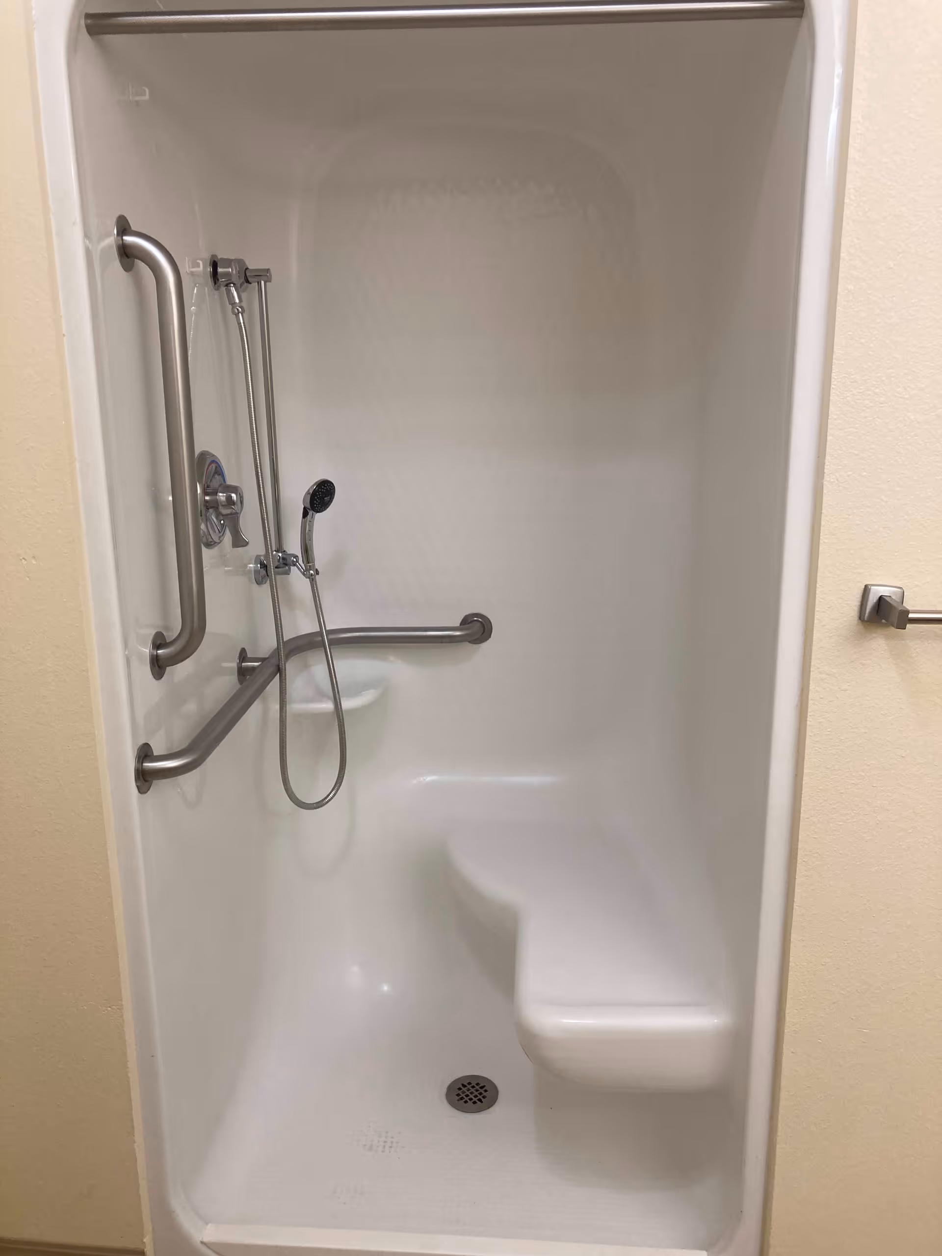 A white shower stall with built-in bench seating and multiple stainless steel grab bars. It has a handheld showerhead attached to a flexible hose and a small soap dish. The walls are smooth and the floor has a drain in the center.