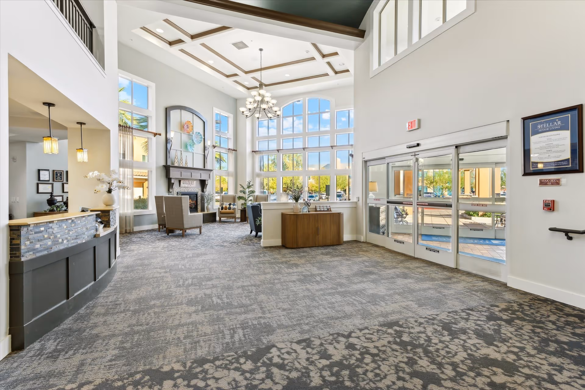 Spacious senior living facility lobby with high ceilings, large windows letting in natural light, a chandelier, a fireplace with decorative items above it, seating area with chairs, a reception desk on the left, and glass automatic entrance doors on the right.